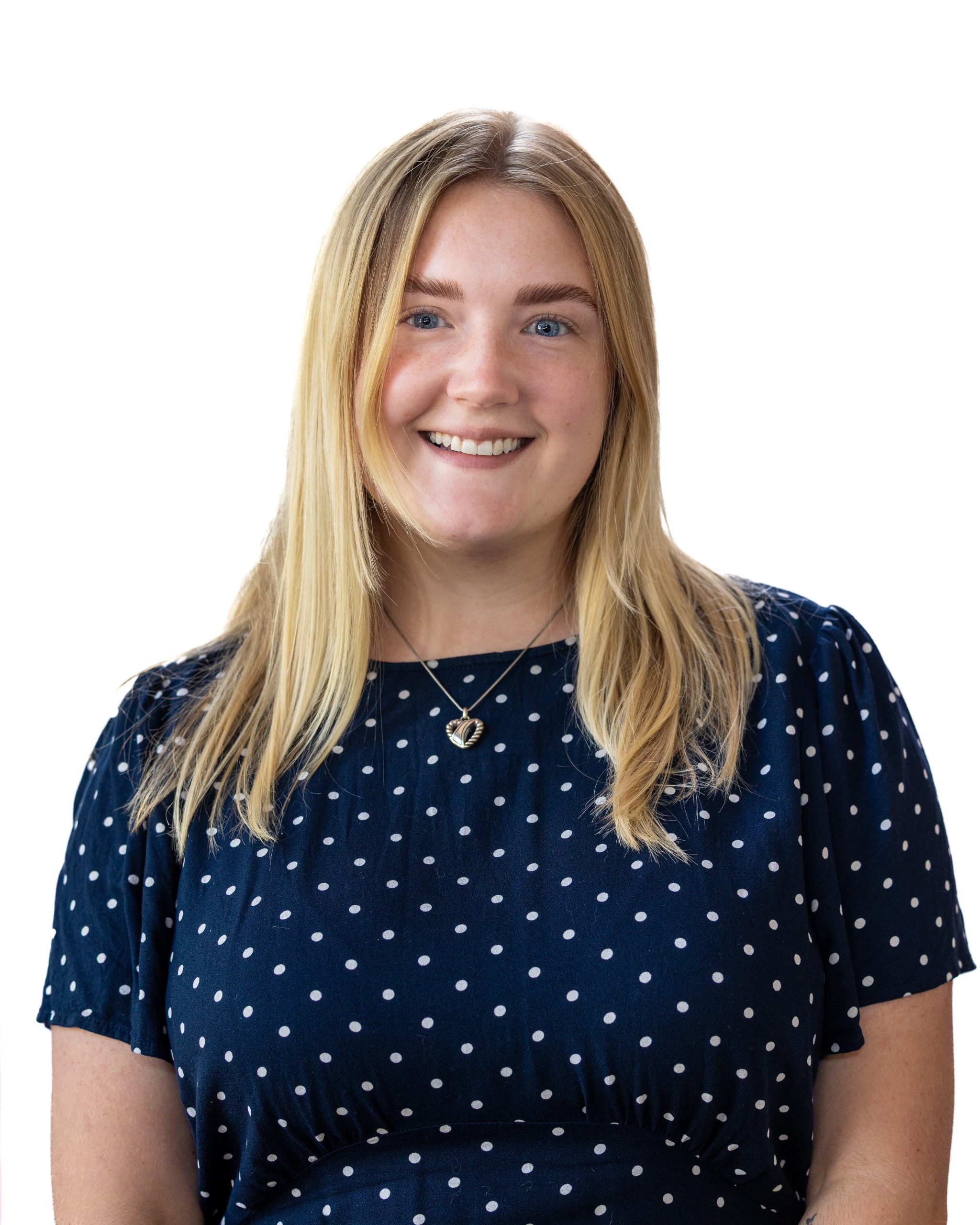 Portrait of a smiling woman with long blonde hair wearing a navy blue polka-dot blouse and heart-shaped necklace, photographed against a white background.